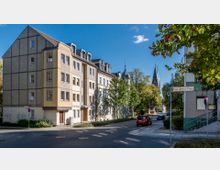 Straßenszene mit mehrstöckigen Wohnhäusern im Vordergrund, einer angrenzenden Straße und vereinzelten Bäumen. Im Hintergrund ist eine Kirche mit spitzem Turm zu sehen; ein Straßenschild zeigt "Karl-Ritter-Platz" an.