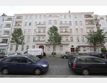 Fünfgeschossiges Wohngebäude mit heller Fassade und mehreren Balkonen, flankiert von Bäumen. Im Vordergrund stehen parkende Autos entlang einer gepflasterten Straße, im Erdgeschoss befindet sich ein kleines Geschäft mit roter Eingangstür.