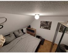 Ein kleines Schlafzimmer mit schräger Decke, ausgestattet mit einem Doppelbett mit grauer karierten Bettwäsche, einer Holzkiste mit schwarzen Schubladen daneben und einem Spiegel an der rechten Wand. Eine Deckenlampe beleuchtet den Raum, und an der Wand hängt ein abstraktes Kunstbild.