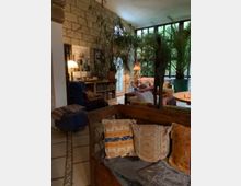 Wohnraum mit gemütlicher Atmosphäre, gestaltet mit Holzmöbeln, farbenfrohen Kissen, großen grünen Pflanzen und Dekorationen wie einer Stehlampe und einer Skulptur. Eine Steinmauer und große Fenster sorgen für natürliches Licht und einen Blick ins Grüne.