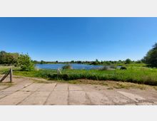 Landschaft mit einem Fluss oder See im Vordergrund, umgeben von Wiesen und Bäumen. Ein unbefestigter Weg sowie ein Metallzaun markieren den Rand des Grasbereichs, während der Himmel klar und blau ist.