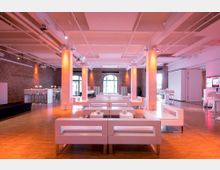 Ein moderner Veranstaltungsraum mit hellen Holzböden, weißen Polstermöbeln und freiliegenden Ziegelwänden. Große Fensterbögen und dezente Beleuchtung schaffen ein offenes und einladendes Ambiente.