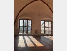 Innenraum mit Kreuzgewölbe und Terrakotta-Fliesenboden, drei hohe Fenster mit Sprossen und Ziegelumrandungen lassen Tageslicht einfallen; Ausblick auf Bäume und historische Architektur im Freien.