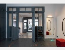 Ein Innenraum mit dunkelgrauen Holzböden und doppelten Glasschiebetüren, die zu einem weiteren Raum mit großen Fenstern führen. An der rechten Wand hängt ein ovaler Spiegel, darunter ein roter Hocker, daneben ein Stuhl und ein Heizkörper. Im Hintergrund ist eine Straße mit parkenden Autos sichtbar.