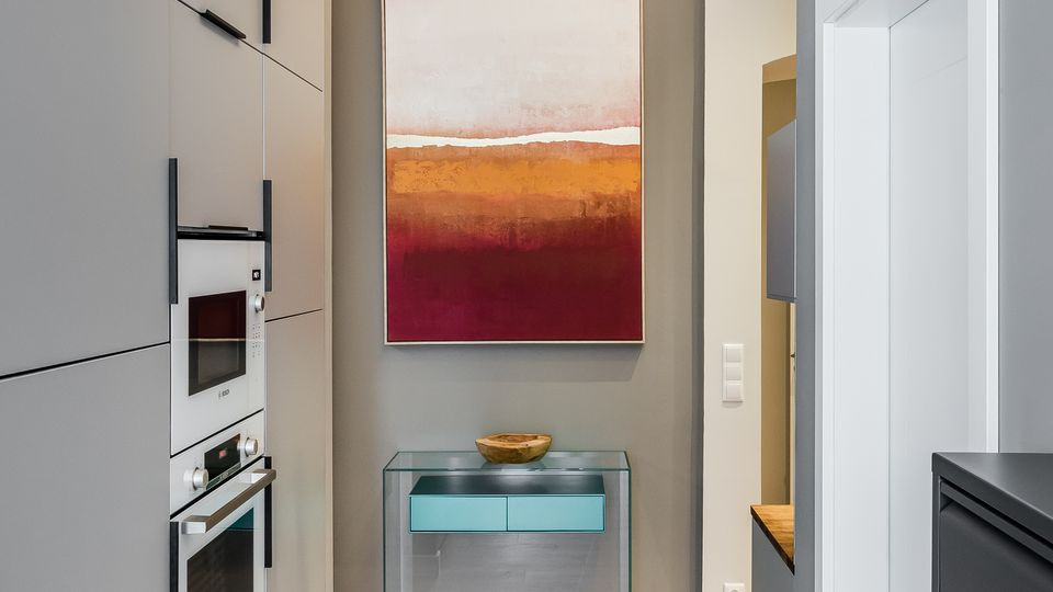 Schmaler, moderner Küchenflur mit hellen Einbauschränken und integrierten Backöfen links, Holzfußboden und grauen Wänden. Am Ende hängt ein abstraktes Bild über einem gläsernen Konsolentisch; die Decke zeigt dekorativen Stuck und eine lineare Deckenleuchte.