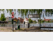 Eingang zu einem Steg mit einem Schild "Herzlich Willkommen am Anleger Neustädtisches Wassertor", umgeben von einem Kopfsteinpflasterweg und überhängenden Baumzweigen. Im Hintergrund befinden sich Gebäude mit roten, weißen und hellen Fassaden sowie ein Kirchturm neben einem Fluss.