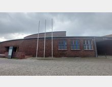 Backsteinbau mit flachem Dach, drei lange Fahnenmasten ohne Fahnen und Fenstern mit farbigen Fensterrahmen in Blau und Rot. Im Vordergrund Pflastersteine und eine breite Gehwegfläche; der Himmel ist bewölkt.