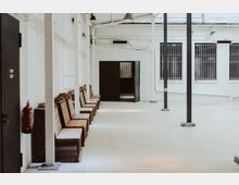 Langer, heller Innenhof mit weißen Wänden, mehreren Sitzbänken aus Holz an der linken Seite und schwarzen Metallpfeilern. Eine offene Tür mit schwarzem Rahmen führt in einen dunkleren Innenraum, daneben sind Fenster mit Gitterstäben zu sehen.