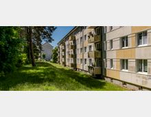 Langer, mehrstöckiger Wohnblock mit beigen Fassadenelementen, vielen Fenstern und vorspringenden Balkonen entlang einer schmalen Grünfläche. Links säumen hohe Bäume und dichtes Gebüsch den Rasenstreifen; im Hintergrund ist ein weiteres Gebäude zu sehen.