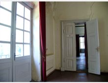 Ein historischer Innenraum mit hohen Doppeltüren und einer großen Fensterfront mit Sprossen. Die Wände sind leicht abgenutzt, und rote Vorhänge hängen neben den Fenstern und Türen.