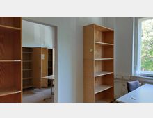 Raum mit einfachen Holzregalen und einem Schreibtisch neben einem Fenster, durch das Tageslicht und der Blick auf Bäume hereinkommen. Im Hintergrund sind weitere Regale und geöffnete Schranktüren sichtbar.