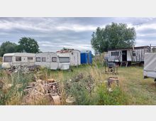 Eine Wiese mit mehreren alten Wohnwagen, einem kleinen hölzernen Schuppen und verstreutem Holz- und Metallmaterial. Im Hintergrund stehen einige Bäume unter bewölktem Himmel.