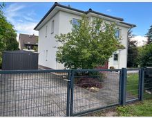 Weißes Einfamilienhaus mit modernem Satteldach und roter Eingangstür, umgeben von einem kleinen Garten mit Bäumen und einem Metallzaun. Im Vordergrund befindet sich eine gepflasterte Einfahrt, links steht ein grauer Geräteschuppen.