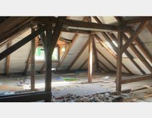 Dachboden mit sichtbaren Holzbalken und abgeflachten Dachschrägen, teils mit Isolierungen verkleidet. Auf dem Boden liegen verstreut Werkmaterialien und Schutt; links und rechts befinden sich Fenster, die Tageslicht hereinlassen.