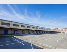 Großer Beton-Verladehof mit einer langen Reihe von blauen Laderampen und Glasfenstern oberhalb, flankiert von leeren Parkflächen. Im Hintergrund ein ziegelrotes Industriegebäude und ein geparkter gelber Lkw.