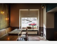 Innenansicht eines gemütlichen Cafés mit Holzmöbeln. Im Vordergrund stehen Holztische und Stühle, im Hintergrund befindet sich ein großes Fenster, durch das eine Straße mit einem roten Auto und grauen Gebäuden sichtbar ist.