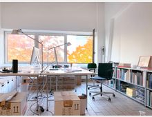 Ein heller Büroraum mit zwei Schreibtischen, auf denen Computer und Schreibtischlampen stehen, umgeben von Kartons und Büchern. Große Fenster im Hintergrund geben den Blick auf herbstbunte Bäume frei, während auf der rechten Seite ein Bücherregal mit Büchern und Ordnern steht.