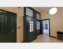 Ein Innenraum mit beige gestrichenen Wänden und dunkelgrünen Holztüren, die mit Glasfeldern versehen sind. Die Türen sind von einem hölzernen Rahmen mit zusätzlichen Glasfenstern umgeben, und eine Tür führt in einen angrenzenden Raum. An der Wand befinden sich ein Brandschalter, ein Lichtschalter und ein Hinweisschild.