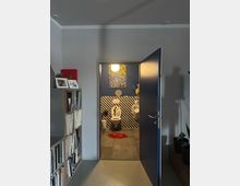 Blick durch eine geöffnete Tür in ein Badezimmer mit gestreifter Wand in Schwarz-Weiß-Optik, einer Toilette, einem Urinal und einer roten, lippenförmigen Matte. Im Vordergrund links ein Regal mit Büchern und Fotos, rechts einige Pflanzen.