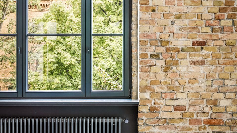 Innenraum mit großer, dunkel gerahmter Fensterfront und Blick auf grüne Bäume; darunter ein breiter Heizkörper. Rechts eine freiliegende Ziegelwand und ein glatter Betonboden im Vordergrund.