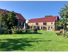 Weitläufiger Garten mit grünem Rasen, Blumenbeeten und Gartenmöbeln, umgeben von zwei Backsteingebäuden mit roten Ziegeldächern; links ein kleineres Haus mit Fensterläden, rechts eine größere Scheune mit Holz- und Glastüren.