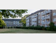 Wohngebiet mit mehrstöckigen Wohngebäuden in schlichtem, hellbraunem Putzstil, jeweils mit Balkonen. Im Vordergrund befindet sich eine gepflegte Grünfläche mit Hecken und vereinzelten Bäumen, umgeben von einer ruhigen Wohnumgebung.