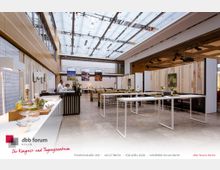 Ein modernes Tagungs- und Veranstaltungszentrum mit hellem, offenem Raum, einer Glasdecke für natürliches Licht, und minimalistischen Holzelementen an den Wänden. Der Raum ist mit hohen weißen Tischen, Sitzgelegenheiten und dekorativen Pflanzen eingerichtet.