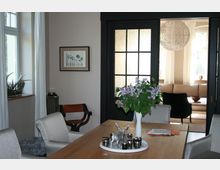 Esszimmer mit einem Holztisch, umgeben von gepolsterten Stühlen, auf dem ein Tablett mit Dekorationen und ein Blumenstrauß steht. Im Hintergrund führt eine schwarze Glastür in ein helles Wohnzimmer mit großen Fenstern und einem Sofa.