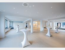 Moderner Veranstaltungsraum mit hellen Wänden und einem perforierten Deckensystem mit eingebauten LED-Leuchten. Der Raum verfügt über hohe Fenster an der linken Seite, mehrere Stehtische mit weißen Tischhussen und Blumendekoration sowie eine offene Küchenzeile im Hintergrund.