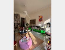 Helles Wohnzimmer mit Holzdielenboden und einer Einrichtung im Retro-Stil, bestehend aus einem grünen modularen Sideboard mit Musikanlage, einer Regalwand voller Schallplatten, einem Couchtisch und einem lilafarbenen Sitzsack. Im Raum befinden sich zudem Dekorationen, bunte Poster und Pflanzen, die durch Sonnenlicht hervorgehoben werden.