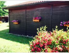 Holzgebäude mit dunkler Fassade, dekoriert mit hängenden Blumenkästen voller bunter Blumen; im Vordergrund ein blühender Strauch mit roten Blüten, umgeben von einer Rasenfläche bei sonnigem Wetter.