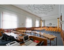 Großer Sitzungssaal mit hölzernen Wänden und Stuckdecke. Der Raum enthält lange Tische mit Computern und Telefonen im Vordergrund sowie zahlreiche Stuhlreihen für Zuschauer oder Teilnehmer, die in Richtung einer Wand mit dekorativen Säulen und einem Wandrelief ausgerichtet sind.