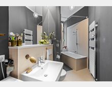 Modernes Badezimmer mit einer Badewanne, einem Waschbecken und einem wandmontierten Handtuchheizkörper. Das Design kombiniert helle Fliesen mit dunklen Wänden, dekoriert mit Pflanzen und Pflegeutensilien.