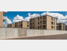 Moderne Wohnanlage mit mehreren rechteckigen, beige verputzten Wohngebäuden, die Rollläden und vereinzelte Balkone besitzen. Im Vordergrund verläuft eine graue Betonmauer entlang einer asphaltierten Straße unter einem Himmel mit weißen Wolken.