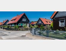 Straßenszene in einem Wohngebiet mit mehreren freistehenden Einfamilienhäusern aus dunklem Holz und roten Ziegeldächern, umgeben von gepflegten Gärten und Zäunen. Im Hintergrund sind weitere Häuser und ein Wald zu sehen, bei klarem, blauem Himmel.