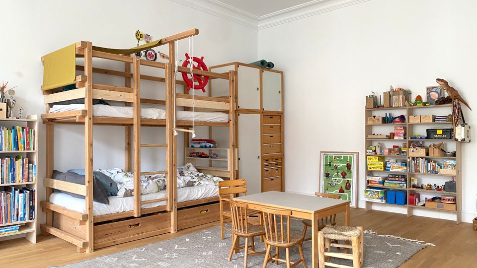 Helles Kinderzimmer mit hohen weißen Wänden und Holzfußboden, ausgestattet mit einem großen Etagenbett aus Holz mit Leiter, Stauraum und Hängeseil. In der Raummitte steht ein kleiner Tisch mit Stühlen auf einem grauen Teppich; an den Wänden stehen Regale und Schränke mit Büchern und Spielzeug.