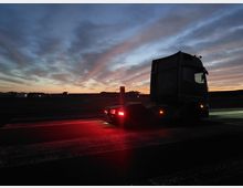 Ein Sattelschlepper steht bei Dämmerung auf einer asphaltierten Straße. Im Hintergrund sind ein heller Horizont, Gebäude und dunkel silhouettierte Landschaft zu erkennen, während der Himmel in Blau- und Orangetönen gefärbt ist.
