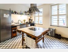 Helle Gemeinschaftsküche mit schwarz-weißen Schachbrettfliesen, Edelstahl-Kühlschrank und Küchenzeile mit Dunstabzugshaube. In der Mitte steht ein großer Holzarbeitstisch mit integriertem Kochfeld; große Fenster sorgen für viel Tageslicht.