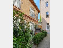 Kleiner Innenhof mit gepflastertem Weg, umgeben von einer gelben Backsteinfassade mit großen, weiß gerahmten Fenstern und Balkongeländern. Im Vordergrund befinden sich grüne Büsche, ein Strandkorb und ein kleiner Holzterrassenbereich.