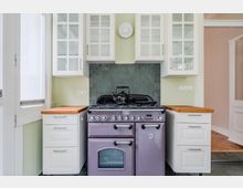 Helle Wohnküche mit großem Gasherd und Backofen in der Mitte, flankiert von weißen Unterschränken mit Holzarbeitsplatten. Darüber hängen weiße Hängeschränke mit Glastüren; die Rückwand ist mit dunkelgrauen Fliesen gestaltet, links ein hohes Fenster und rechts ein Türdurchgang.