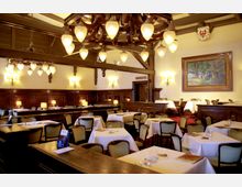 Innenansicht eines traditionellen Restaurants mit Holzvertäfelung und dekorativen Deckenbalken. Der Raum ist mit weißen Tischdecken, vintage Stühlen, Wandleuchten und einem großen Kronleuchter ausgestattet, im Hintergrund hängt ein großes Gemälde an der Wand.