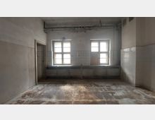 Ein leeres, verfallenes Zimmer mit zwei großen Fenstern, abgenutztem Holzboden und gefliesten Wänden bis zur halben Höhe. An der Wand unter den Fenstern befinden sich alte Metallspülbecken, und über den Fenstern sind Rohre sichtbar.