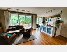 Wohnbereich mit offener Küche und Zugang zu einer Terrasse. Der Raum hat Parkettboden, eine Sitzecke mit Ledermöbeln und einen Holztisch, sowie eine weiße Einbauküche mit Hängelampen; große Fenster bieten Blick auf grüne Bäume.