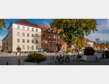 Städtischer Platz mit gepflastertem Boden, umgeben von historischen Gebäuden, darunter ein rotes Backsteingebäude und ein beiges Haus. Im Vordergrund stehen Fahrräder an Fahrradständern sowie große Bäume und Pflanzkübel.