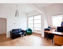 Heller Büroraum mit weißer Decke und schrägen Dachflächen, großen Fenstern und hellem Holzfußboden. Links stehen ein schwarzes Sofa und ein grüner Sessel mit kleinem Tisch, rechts ein großer Schreibtisch mit Computer.