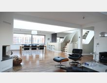 Moderner Wohnbereich mit offener Gestaltung, einer Holztreppe mit geschwungenem Geländer, großem Esstisch mit schwarzen Stühlen, Parkettboden und einem Kamin auf der linken Seite. Große Fenster lassen viel Tageslicht herein, und eine Lounge-Ecke mit Ledersessel und Beistelltisch befindet sich im Vordergrund.