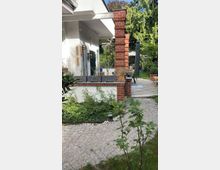 Wohnhaus mit weißer Fassade und roten Backsteinelementen an einem Pfeiler. Im Vordergrund ein gepflasterter Weg mit umliegender Vegetation, dahinter eine Terrasse mit Pflanzen in Töpfen und Gartenmöbeln im Grünen.