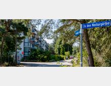 Straßenszene mit einem Wohngebäude auf der linken Seite, das von Bäumen und viel Grün umgeben ist. Im Vordergrund steht ein Straßenschild mit der Aufschrift „Zu den Neckargärten“.