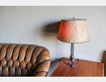 Braunes, gestepptes Ledersofa neben einem Holztisch mit einer Tischlampe; die Lampe hat einen beige strukturierten Schirm und einen dekorativen, gedrehten Metallfuß.