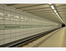 U-Bahnhof mit glatten, weiß gefliesten Wänden und einem durchgehenden grünen Streifen mit der Aufschrift „Strausberger Platz“. Der Bahnsteig ist leer, das Gleis verläuft parallel zur gefliesten Wand, und die Decke ist mit gleichmäßigen Leuchtstofflampen beleuchtet.